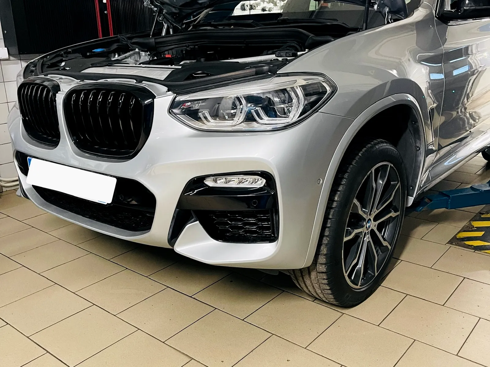Sidabrinis BMW X3 su atidarytu variklio dangčiu ant keltuvo Autvadita servise