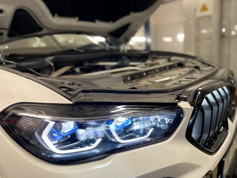 Baltas BMW su atidarytu variklio dangčiu ir įjungtais lazeriniais žibintais serviso bokse