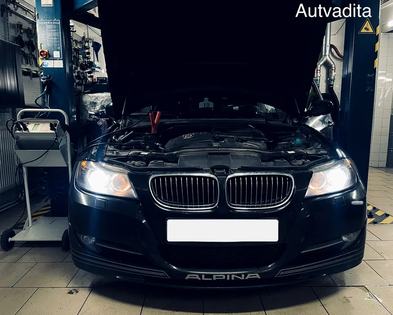 Juodas BMW Alpina ant keltuvo su atidarytu dangčiu ir prijungtu akumuliatoriaus įkrovikliu Autvadita servise