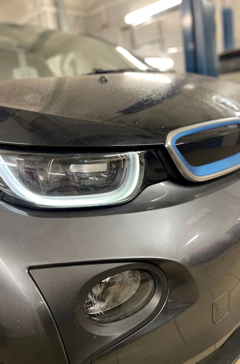BMW i3 priekinis žibintas ir grotelės su mėlynais apvadais iš arti serviso patalpoje