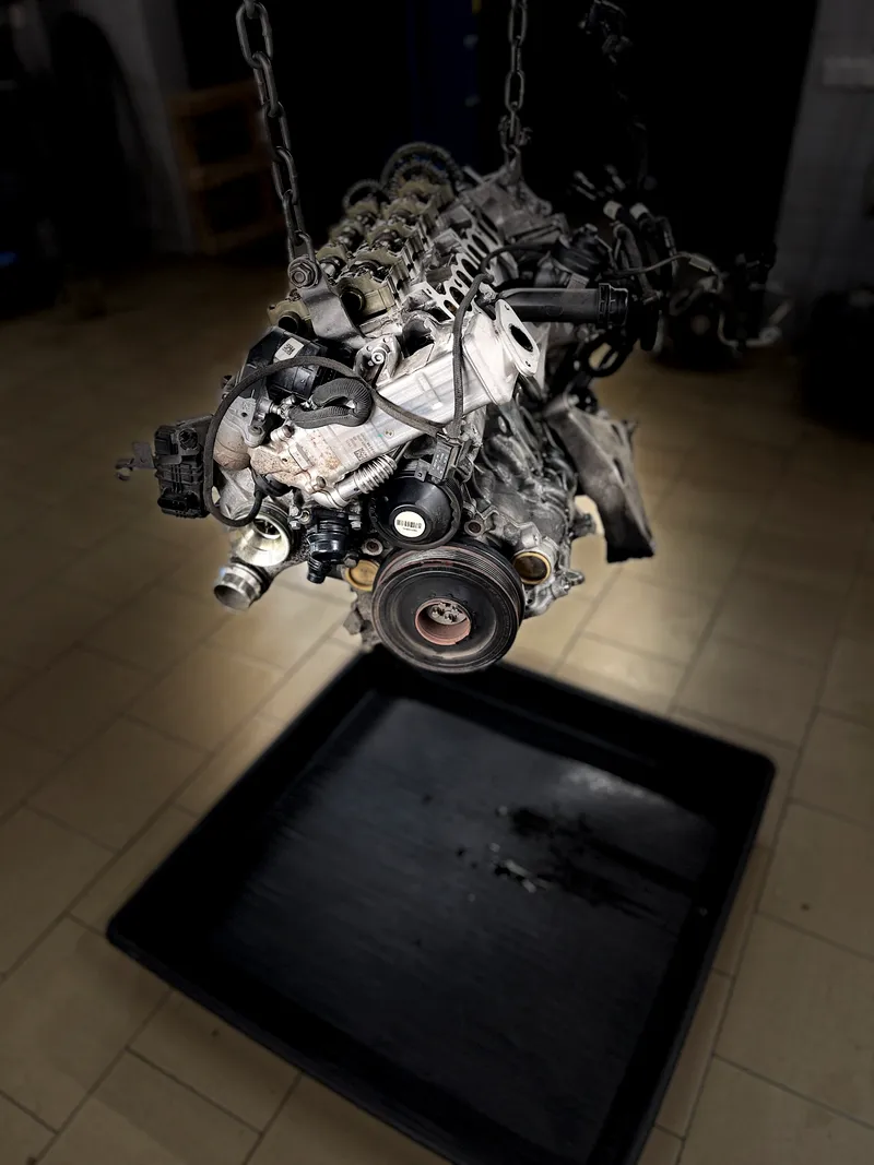 Iš automobilio išimtas BMW variklis, pakabintas ant keltuvo grandinių virš surinkimo padėklo