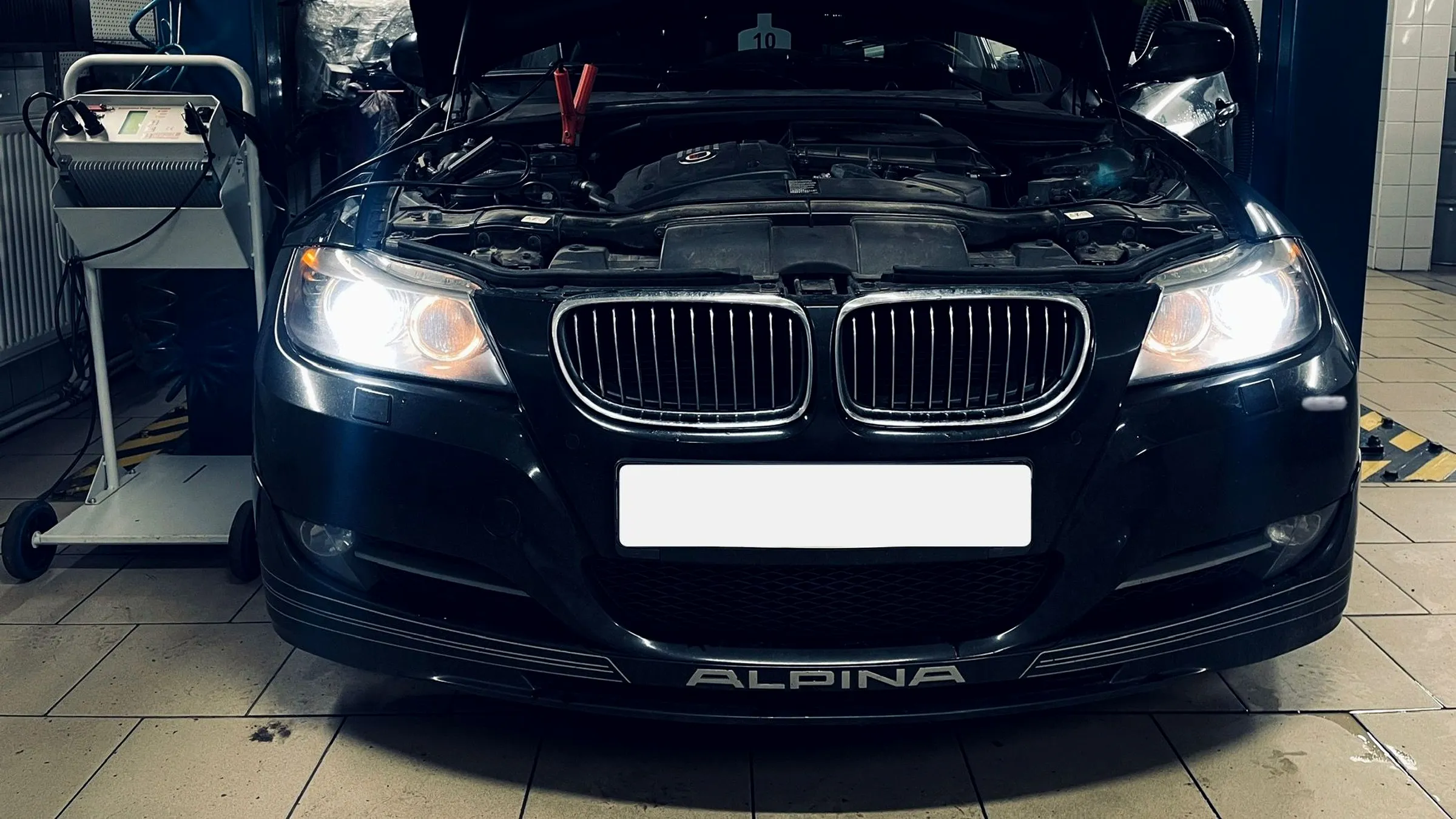 Juodas BMW Alpina ant keltuvo su atidarytu variklio dangčiu ir prijungtu akumuliatoriaus įkrovikliu Autvadita servise