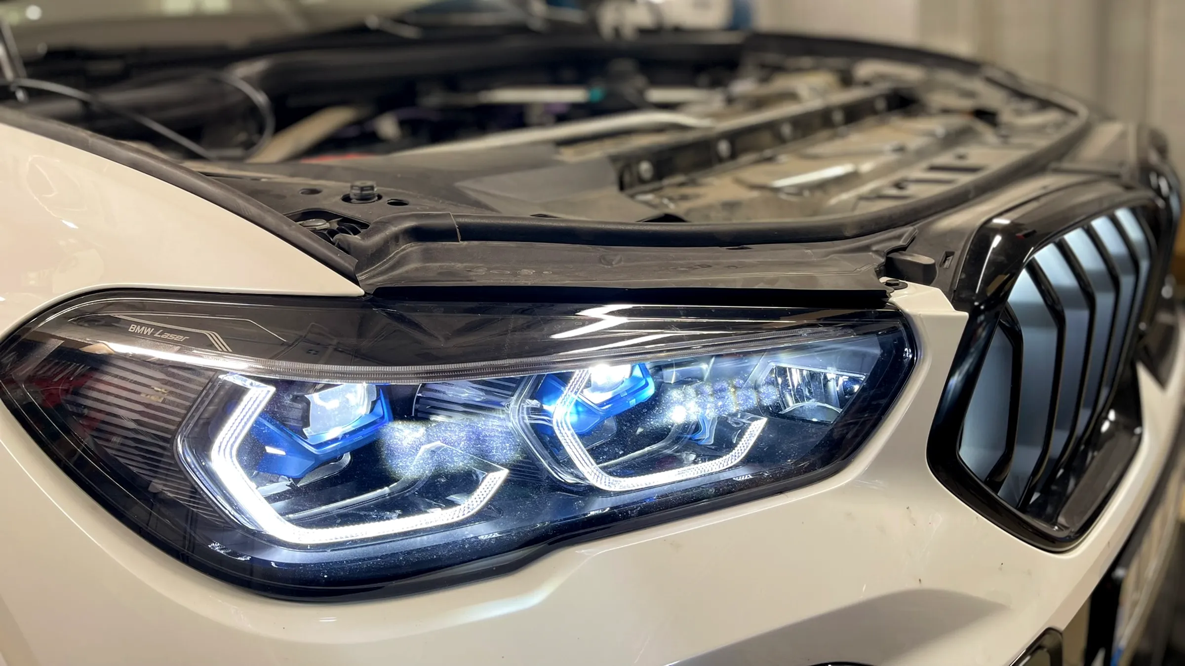 Baltas BMW su atidarytu kapotu ir įjungtais lazeriniais žibintais servise